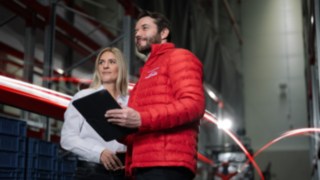 A man in a red jacket from Linde MH and a woman in a white shirt stand in a warehouse and look into the distance.