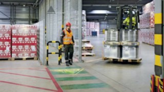 A pedestrian crosses the path of a turning forklift truck and a warning symbol appears on the hall floor in front of them.